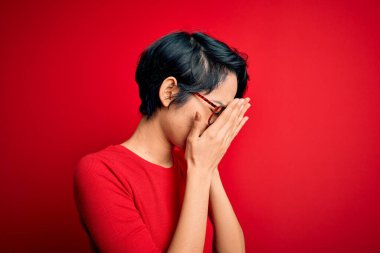 Genç, güzel Asyalı bir kız, günlük tişört ve gözlük takıyor. İzole edilmiş kırmızı arka plan üzerinde. Üzücü bir yüz ifadesi var. Depresyon kavramı.