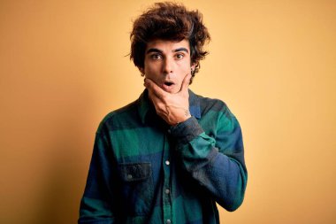 Sarı arka plan üzerinde duran sıradan tişörtlü genç yakışıklı adam elleri çenesinde, inanamama, şaşkınlık ve şaşkınlık dolu bir ifade takınıyor.