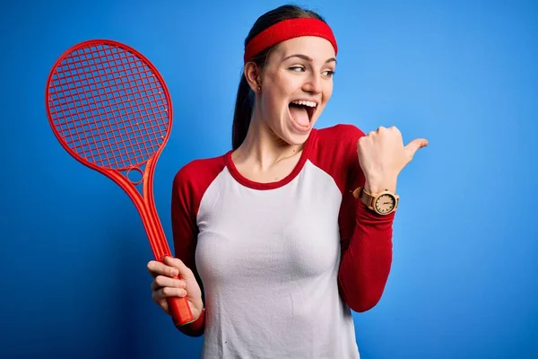 若いです美しいです赤毛スポーツ女性プレイテニスラケットを使用して上の青の背景ポインティングと表示しますとともに親指で側まで幸せな顔笑顔 — ストック写真
