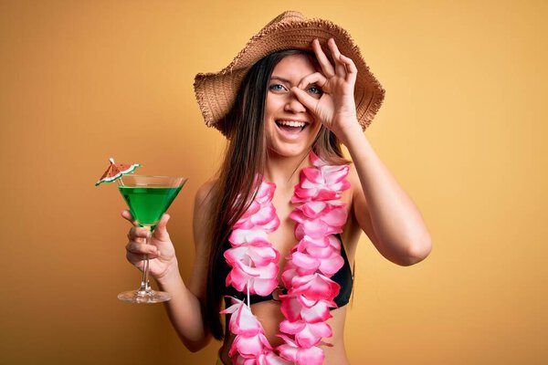 Beautiful woman with blue eyes on vacation wearing bikini and hawaiian lei drinking cocktail with happy face smiling doing ok sign with hand on eye looking through fingers