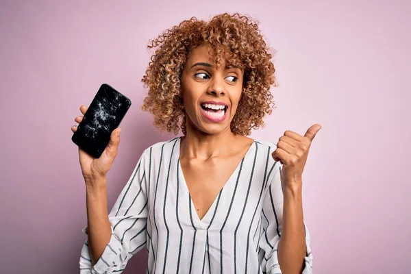 Young african american curly woman holding broken smartphone showing craked screen pointing and showing with thumb up to the side with happy face smiling
