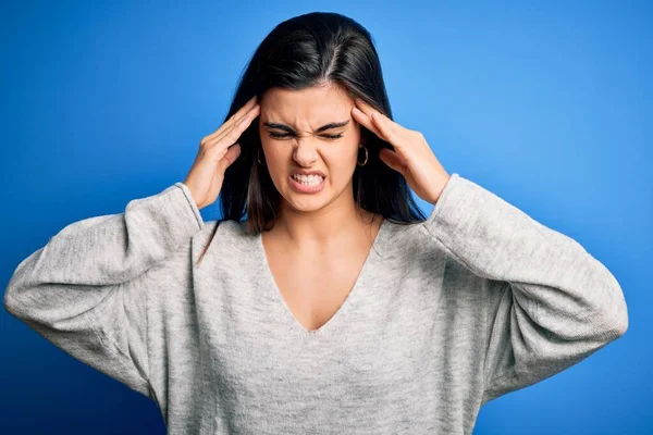 ストレスのために頭の痛みのために頭の上に手で青の背景に立ってカジュアルなセーターを着て若い美しいブルネットの女性 片頭痛に苦しむ — ストック写真