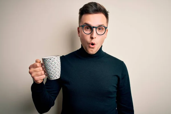 Young handsome caucasian man wearing glasses drinking a cup of hot coffee scared in shock with a surprise face, afraid and excited with fear expression