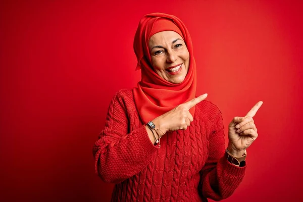 Middle age woman wearing traditional muslim hijab standing over isolated red background smiling and looking at the camera pointing with two hands and fingers to the side.