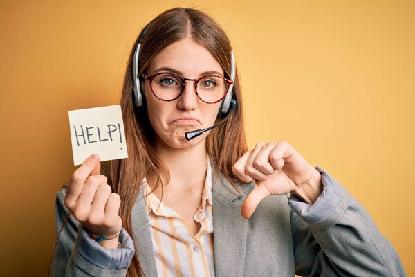 Молодая рыжая агент колл-центра женщина перегружена работой с помощью гарнитуры, держа помощь остаток с сердитым лицом, отрицательный знак, показывающий неприязнь с опущенными пальцами, отвержение концепции
