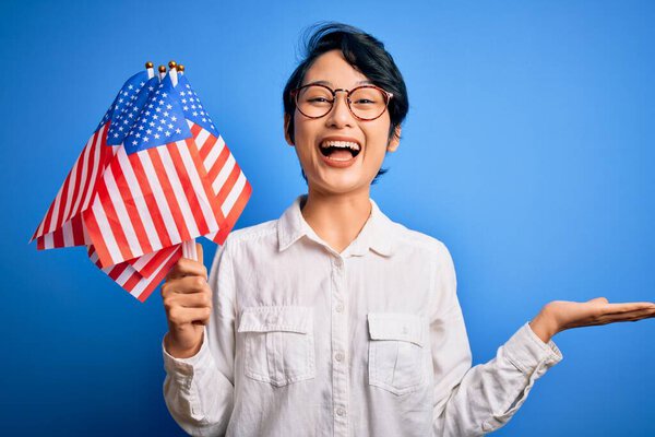 Young beautiful asian patriotic girl holding united states flags celebrating independence day very happy and excited, winner expression celebrating victory screaming with big smile and raised hands