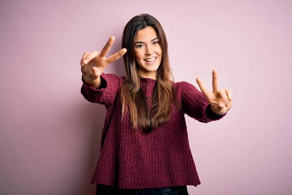 孤立したピンクの背景の上にカジュアルなセーターを着ている若い美しい女の子は 勝利のサインをして両手の指を見せて舌で笑っています 第二番 — ストック写真