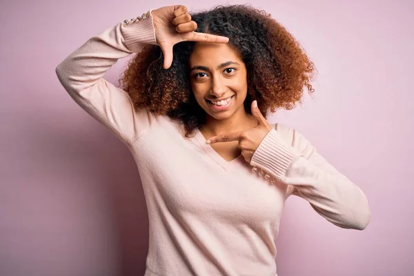 ピンクの背景にカジュアルなセーターを着たアフロヘアの若いアフリカ系アメリカ人女性は 幸せな顔をした手や指でフレームを作り笑顔 創造性と写真の概念 — ストック写真