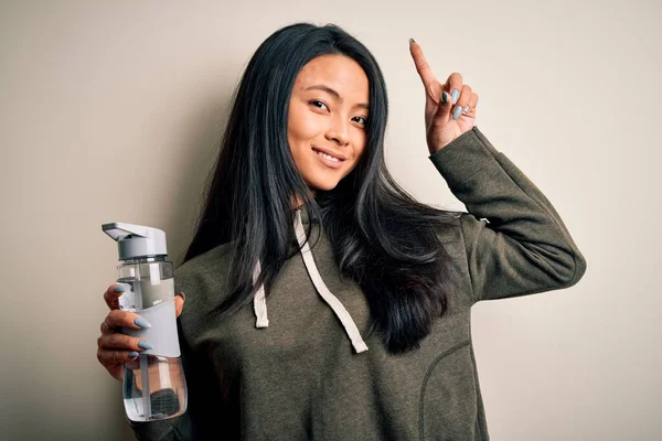 Young beautiful chinese sportswoman holding bottle of water over isolated white background surprised with an idea or question pointing finger with happy face, number one