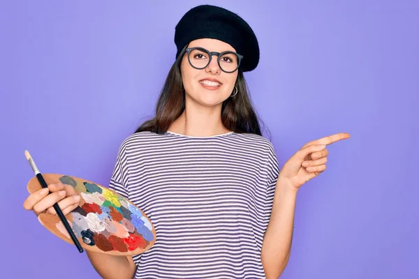 筆とパレットを使用してファッションベレー帽の絵画を身に着けている若い美しい芸術家の女の子側に手と指で非常に幸せなポインティング — ストック写真