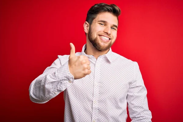 髭と青い目をした若いブロンドのビジネスマンは 赤い背景にエレガントなシャツを着て 手で幸せな親指を立ててジェスチャーをします 成功を示すカメラを見て表現を承認する — ストック写真