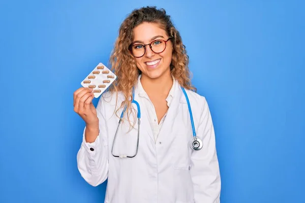 Young beautiful blonde doctor woman with blue eyes wearing stethoscope holding pills with a happy face standing and smiling with a confident smile showing teeth