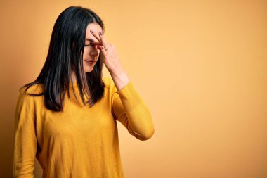 Mavi gözlü genç esmer kadın sarı arka planda rahat süveter giyiyor. Burnu ve gözleri ovuşturuyor, yorgunluk ve baş ağrısı çekiyor. Stres ve hüsran kavramı.