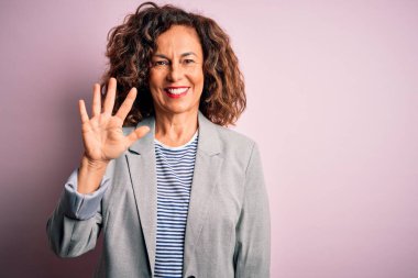 Orta yaşlı, güzel bir iş kadını zarif bir ceket giyip pembe arka planda beş numaralı parmaklarıyla işaret ederken kendinden emin ve mutlu bir şekilde gülümsüyor..