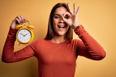 Genç, güzel esmer kadın, sarı arka planda alarmlı bir saat tutuyor. Mutlu bir yüzü var. Gülümsüyor.