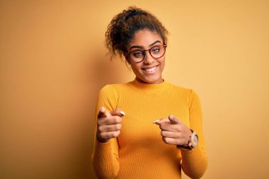Sarı arka planda süveter ve gözlük takan güzel Afro-Amerikan kızı mutlu ve komik yüzlü parmaklarını kameraya doğrultuyor. İyi enerji ve titreşimler.