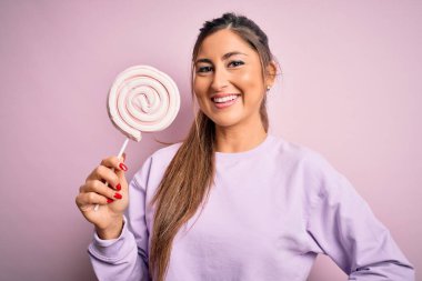 Genç, güzel esmer kadın pembe arka planda tatlı şekerler yiyor. Mutlu bir yüzü var ve gülümsüyor. Kendine güvenen bir gülümsemeyle dişlerini gösteriyor.