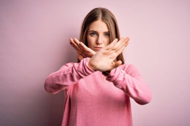 Genç, güzel, kızıl saçlı bir kadın pembe arka planda günlük süveter giyiyor. Reddedilmiş yüz ifadesi. Kollarını kavuşturup negatif işaret veriyor.