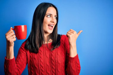 Mavi gözlü genç bir kadın izole edilmiş arka planda duran bir fincan kahve içiyor ve gülümseyen bir gülümsemeyle baş parmağıyla işaret ediyor.