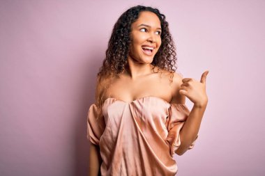 Kıvırcık saçlı, rahat tişörtlü pembe arka planda duran mutlu yüzüyle gülümseyen ve baş parmağıyla yüzünü gösteren güzel bir kadın..