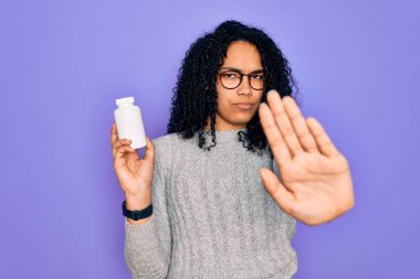 Genç Afro-Amerikan kıvırcık kadın elinde haplarla mor arka planda duruyor. Açık elle dur işareti yapıyor. Ciddi ve kendinden emin bir ifadeyle.