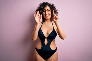 Genç, güzel bir Arap kadın tatilde. Pembe arka planda mayo ve güneş gözlüğü takıyor. Altıncı parmaklarıyla işaret ediyor ve gülümsüyor..