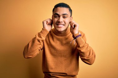 İzole edilmiş sarı arka planda dikilen sıradan süveter giyen genç Brezilyalı adam parmaklarıyla kulaklarını çekerek gülüyor, komik bir jest. Seçme sorunu