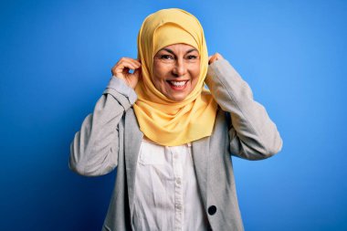 Orta yaşlı esmer iş kadını, geleneksel Müslüman tesettüre bürünmüş, mavi arka planda gülümseyen, parmaklarıyla kulaklarını çeken, komik bir jest. Seçme sorunu