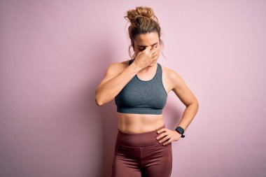 Genç ve güzel sarışın sporcu kadın spor yapıyor. Pembe arka planda spor kıyafetleri giyiyor. Burnunu ve gözlerini ovuşturup yorgunluk ve baş ağrısı çekiyor. Stres ve hüsran kavramı.