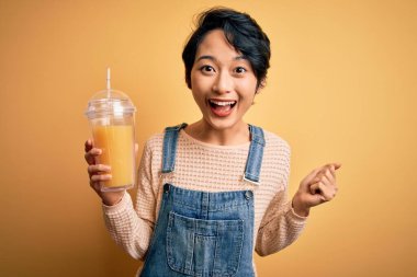 Genç, güzel Çinli bir kadın izole edilmiş sarı arka planda sağlıklı portakal suyu içiyor. Gururla bağırıyor, zaferi kutluyor ve başarıyı kutluyor. Çok heyecanlı, coşkulu bir duygu.