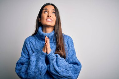 Genç, güzel esmer kadın beyaz arka plan üzerine balıkçı yaka kazak giyip elleri bağlı yalvarıyor ve dua ediyor. Yüzünde umut ifadesi var. Çok duygusal ve endişeli. Yalvarıyorum..