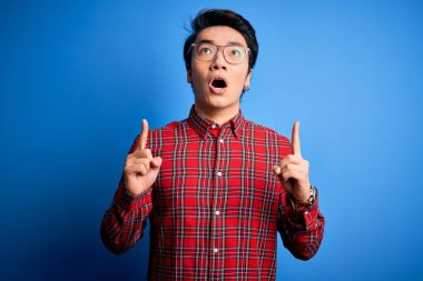 Mavi arka planda günlük gömlek ve gözlük takan genç yakışıklı Çinli adam şaşırmış ve şaşırmış bir şekilde parmaklarıyla işaret ediyor ve kollarını kaldırıyordu..