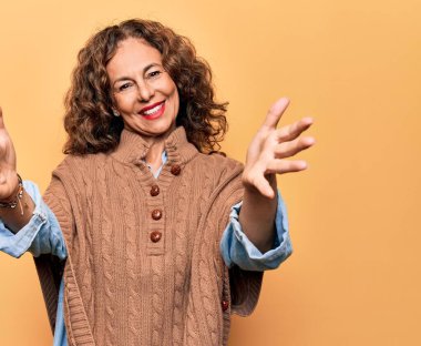 SarıImak için kollarını açıp gülümseyen, izole edilmiş sarı arka plan üzerinde duran orta yaşlı, güzel bir kadın. Mutluluğu kucaklayan neşeli bir ifade..