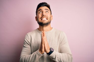 Soyutlanmış pembe arka plan üzerinde duran sıradan bir süveter giyen genç yakışıklı adam elleri bağlı yalvarıyor ve dua ediyor. Yüzünde umut ifadesi var. Çok duygusal ve endişeli. Yalvarıyorum..