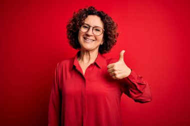 Orta yaşlı güzel kıvırcık saçlı kadın kırmızı arka planda günlük gömlek ve gözlük takıyor. Elle mutlu bir jest yapıyor. Kameraya bakıp başarıyı gösteren ifade onaylanıyor.