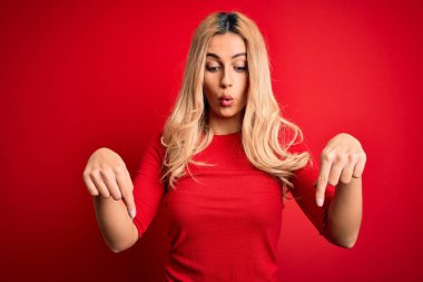 Genç ve güzel sarışın bir kadın, günlük tişört giyiyor, kırmızı arka plan üzerinde duruyor, parmaklarını göstererek reklamını yapıyor, şaşırmış yüzünü ve ağzını açıyor.