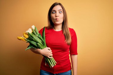 Sarı arka planda romantik lale çiçekleri tutan genç sarışın kadın dudaklı balık suratı yapıyor, çılgınca ve komik bir jest. Komik ifade.