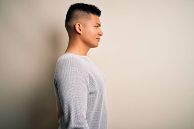 Genç, yakışıklı, günlük süveter giyen, beyaz arka planda duran, kendine güvenen, doğal yüzüyle poz veren Latin bir adam..