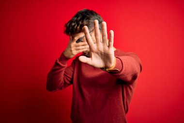Sakallı, gözlüklü ve süveterli genç bir adam kırmızı arka planda dikiliyor gözleri ellerle kaplıyor ve üzgün ve korku ifadeleriyle jest yapıyor. Utanç verici ve olumsuz bir kavram..