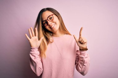 Mavi gözlü güzel sarışın kadın pembe arka planda süveter ve gözlük takıyor ve yedinci parmaklarıyla işaret ederken kendinden emin ve mutlu gülümsüyor..