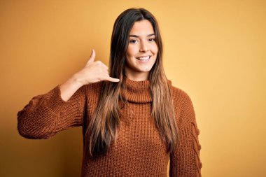 İzole edilmiş sarı arka planda dikilen sıradan süveter giyen genç ve güzel bir kız telefonla el ve parmaklarıyla telefonla konuşuyor gibi gülümsüyor. İletişim kavramları.