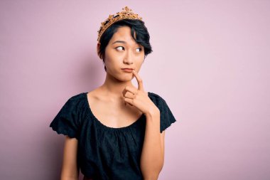 Genç, güzel Çinli bir kadın, kralın altın tacını takmış pembe arka planda, elleri çenesinde soru sorarken, düşünceli bir ifade takınıyor. Düşünceli bir yüzle gülümsüyor. Şüphe kavramı.