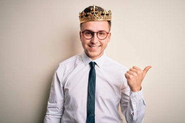 İzole edilmiş arka planda altın taç takan yakışıklı beyaz iş adamı gülümsüyor ve başparmağıyla yüzünü yukarı kaldırıyor..