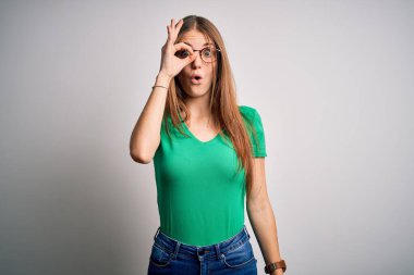Genç, güzel, kızıl saçlı, sıradan yeşil tişörtlü ve gözlüklü bir kadın beyaz arka plan üzerinde şaşırmış yüz ifadesiyle şaşırmış, parmaklarından bakan bir jest yapıyor. İnançsız ifade.