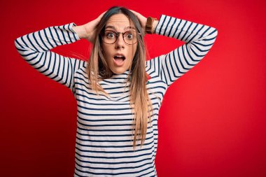 Genç, sarışın, mavi gözlü, gözlüklü, kırmızı arka planda duran, elleri kafasında çılgın, korkmuş ve ağzı açık olan, şoktan korkan bir kadın.