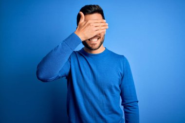 Sakallı, sıradan süveter giyen, mavi arka planda gözlük takan, gülümseyen ve gülen genç yakışıklı bir adam. Kör kavram.