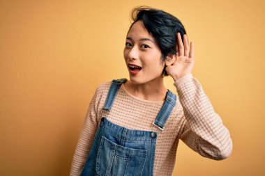 İzole edilmiş sarı arka planda rahat kot pantolon giyen genç, güzel Asyalı bir kız kulağından girip kulağından çıkan dedikoduları ya da dedikoduları dinliyor. Sağırlık kavramı.