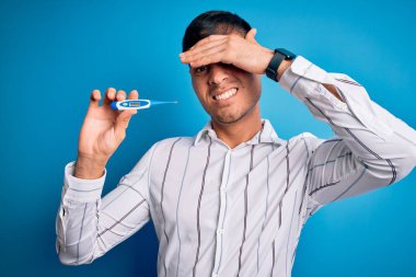 Tıbbi termometreyi mavi izole arkaplanda tutan genç bir İspanyol, elleri başının üstünde, utanç ve şaşkınlık dolu, sinirli ve hayal kırıklığına uğramış bir halde. Hata için korku ve üzüntü.