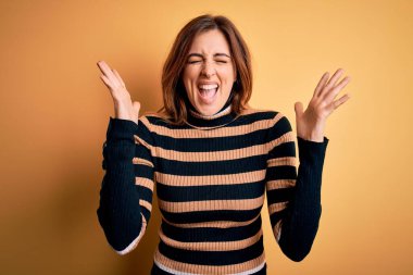 Sarı arka plan üzerine çizgili balıkçı yaka kazak giyen genç, güzel esmer kadın kollarını kaldırmış ve gözlerini kapatmış heyecanla haykırarak başarıyı kutluyor. Kazanan konsept