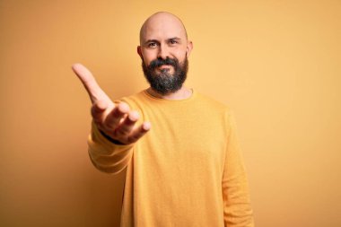 Sarışın, sakallı, sıradan bir süveter giyen yakışıklı adam sarı arka planda durup dostça gülümseyerek selamlaşma ve selamlaşma sunuyor. Başarılı bir iş..